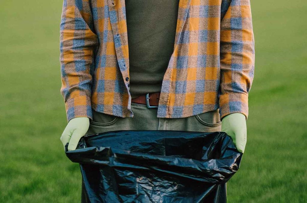 Lower angle image of a man with protective gloves holding a black garbage bag.